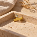 A minimalist gold ring featuring a geometric bar and square stone on a clean white background